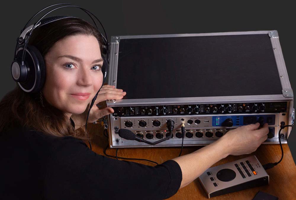 Frau mit AKG-Kopfhörern arbeitet an einem professionellen Audio-Interface und Mischpult, während sie in die Kamera lächelt.